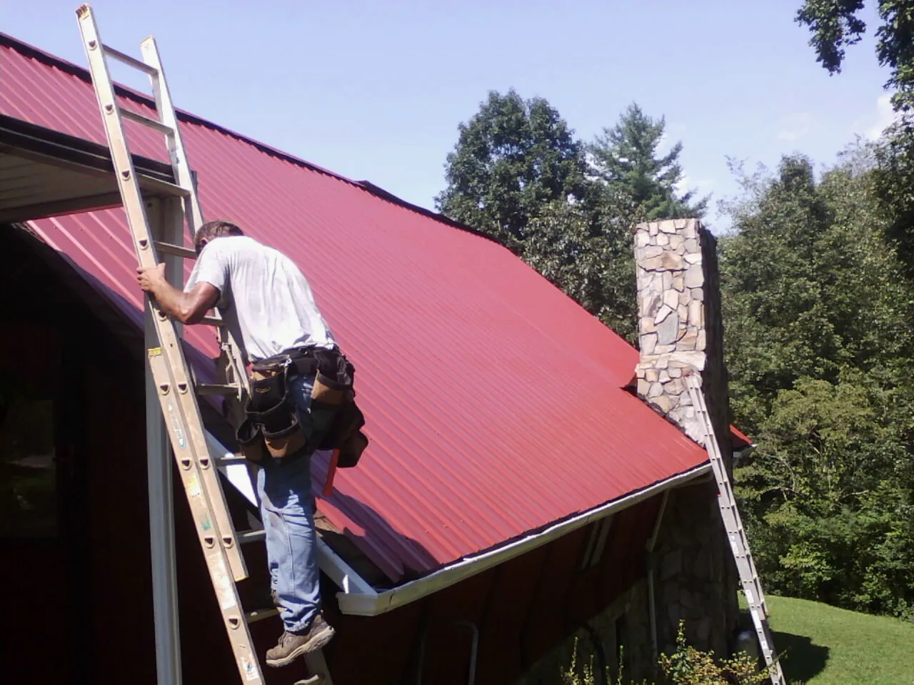 Roofer on a ladder beside a red metal roof and stone chimney — Soffit & Fascia Repair by Roofing Brothers in Mount Arlington